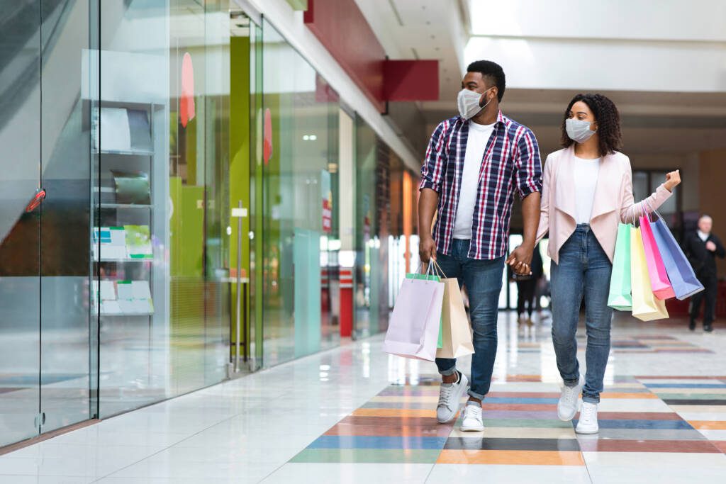 Pandemic Shopping. Black couple wearing protective face medical masks walking with shopper bags in mall, african american spouses making purchases during coronavirus 
