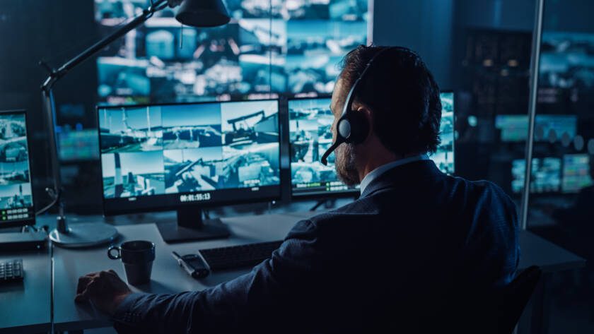 Male Officer Works on a Computer with Surveillance CCTV Video in a Harbour Monitoring Center with Multiple Cameras on a Big Digital Screen
